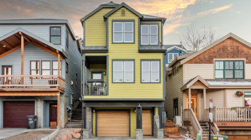 A modern three-story house with a yellow exterior, flanked by two other homes, set against a sunset sky.